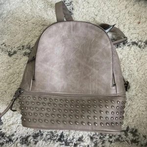 Grå ryggsäck från Co & Coo Fashion - Säljer en snygg grå ryggsäck från Co & Coo Fashion. Väskan är gjord av syntetmaterial och har en cool design med nitar längst ner. Den har justerbara axelremmar och en dragkedja för att hålla dina saker säkra. Perfekt för både vardag och fest!