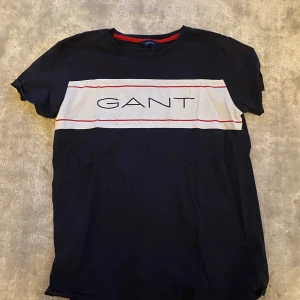 Svart t-shirt från GANT - Säljer en snygg mörk blåa t-shirt från GANT. Den har en vit och röd randig detalj över bröstet med GANT-loggan i mitten. T-shirten är i bomull och har en normal passform. Perfekt för vardagsbruk eller en avslappnad dag ute. Skicket är bra men en liten tråd hänger från den. Passa på att fynda! 😎