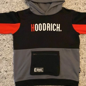 Svart, grå och röd hoodie från Hoodrich - Säljer en snygg hoodie från Hoodrich i färgerna svart, grå och röd. Den har en stor ficka med dragkedja på framsidan och texten 'Hoodrich' tryckt både fram och bak. Perfekt för höst och vinter!