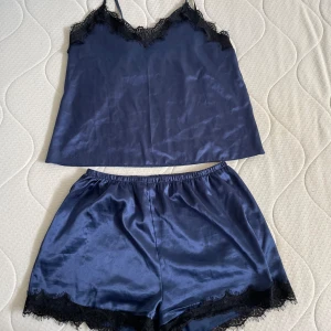Mörkblå satinlinne med spetsdetaljer - Säljer ett elegant mörkblått satinlinne med spetsdetaljer vid halsringningen. Linnet har en lös passform, perfekt för en bekväm och lyxig känsla. Matchande shorts med spetskant ingår.