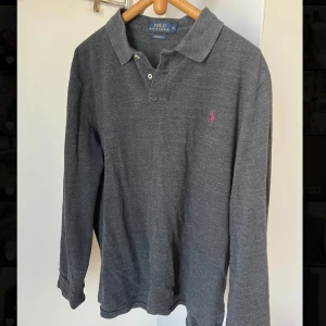 Mörkgrå pikétröja från Ralph Lauren - Säljer en snygg och stilren mörkgrå pikétröja från Ralph Lauren. Tröjan har långa ärmar och en klassisk krage med två knappar. Den är i bra skick och har Ralph Laurens ikoniska logga broderad i vinrött på bröstet. Perfekt för både vardag och lite finare tillfällen. Passar bra till jeans eller chinos. Skicka ett meddelande om du är intresserad! Den är i storlek xl i barn storlek men motsvarar M! Tröjan passar perfekt nu till hösten 🍁