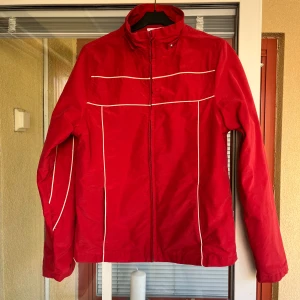 Röd jacka från AMiCA - Säljer en röd jacka från AMiCA i storlek 34. Jackan har en dragkedja framtill och vita detaljer som ger den en sportig look. Den är tillverkad av 78% bomull och 22% nylon, med ett foder av 100% polyester. Avtagbar luva. Perfekt för höst & vår.