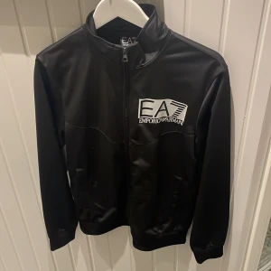 EA7 tröja med dragkedja - Svart Emporio Armani tröja med dragkedja från botten till toppen.  Väldigt fin tröja utan några skador som hål eller fläckar.  Tröjan är i storlek S-M och är i de tunnare materialet.  Gammal modell av EA7 som är väldigt snygg. Köpt från johnells för ett par år sen. 
