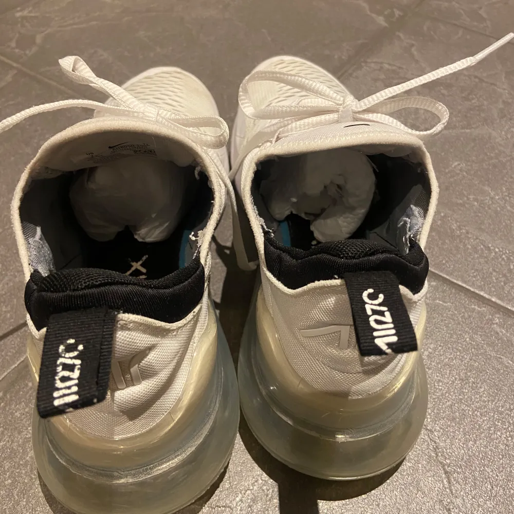 Säljer ett par fräscha vita Nike Air Max 270 sneakers. De har en snygg och modern design med en stor synlig Air-enhet i hälen för extra komfort. Skorna har en vit ovandel med svarta detaljer och snörning. Perfekta för både vardag och träning!. Skor.