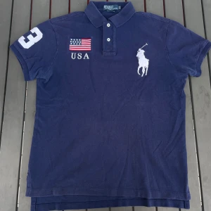 Mörkblå pikétröja från Ralph Lauren - Säljer en snygg mörkblå pikétröja från Ralph Lauren. Den har korta ärmar och en klassisk krage med knappar. På framsidan finns en stor vit broderad logga av en polospelare, samt en USA-flagga och texten 'USA'. På ena ärmen finns siffran '3' broderad. Perfekt för en casual look eller en dag på stan!
