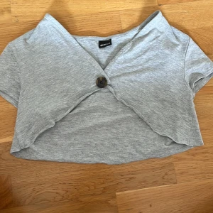 Grå croppad topp från Gina Tricot - Säljer en snygg grå bolero topp från Gina Tricot. Den har en unik omlott-design framtill med en knappdetalj. Toppen är kortärmad och perfekt för sommaren. Materialet känns mjukt och bekvämt, troligen bomull eller en bomullsblandning. Perfekt för en avslappnad men stilren look!