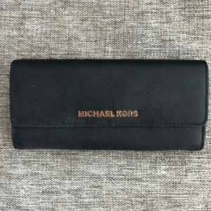 Svart plånbok från Michael Kors - Säljer en stilren svart plånbok från Michael Kors. Den har flera fack för kort och en dragkedja för mynt. Perfekt för att hålla ordning på dina saker med stil. Plånboken är i bra skick och har en elegant design med Michael Kors-loggan i guld på framsidan.