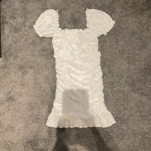 Vit klänning från Gina Tricot - Säljer en superfin vit klänning från Gina Tricot. Den har puffiga kortärmar som kan bäras offshoulder och en söt volangkant längst ner. Klänningen är i ett broderat material och har en tight passform som framhäver figuren. Perfekt för sommarens fester eller en mysig midsommar!