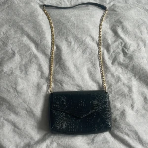Svart kuvertväska från Anna Field - Säljer en snygg svart kuvertväska från Anna Field. Väskan har en elegant krokodilpräglad yta och en stilren design. Den har en guldfärgad kedjerem. Väskan är i nyskick och redo att användas!