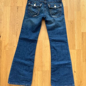 Blå jeansbyxor från True Religion - Säljer ett par snygga blå bootcut jeans från True Religion. Byxorna har coola detaljer som knappar och dragkedja, samt de klassiska True Religion-broderierna på bakfickorna. Står strl 10 men vet inte vad det innebär🫶🏻