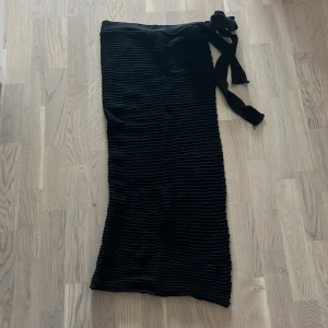 Svart ribbad långkjol med knytband - Säljer en snygg svart strandkjol med ribbad struktur. Kjolen har ett knytband i midjan som ger en fin detalj och gör att den sitter perfekt. kjolen är öppen på ena sidan 