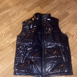 Svart glansig dunväst - Snygg svart glansig dunväst med dragkedja framtill. Västen har två praktiska fickor och en hög krage för extra värme. Perfekt för lo kyligare dagar när du vill hålla stilen.