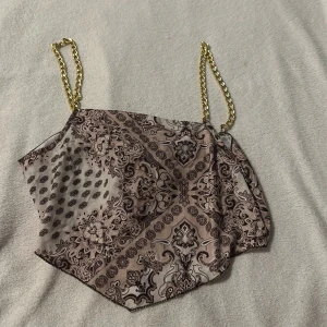 Mönstrad linnetopp med guldkedjor från Amisu - Snygg mönstrad linnetopp från Amisu med guldkedjor som axelband. Toppen har ett unikt mönster i beige och brunt och knyts i ryggen för en perfekt passform. Perfekt för att ge en touch av glamour till din outfit.