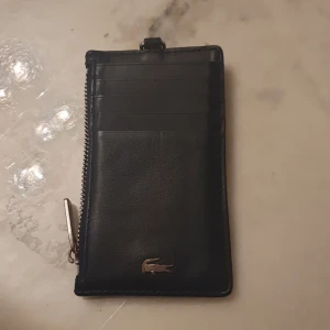 Svart plånbok från Lacoste - Snygg svart plånbok i skinn från Lacoste med flera kortfack och en dragkedja. Perfekt för att hålla ordning på kort och kontanter. Den har en stilren design med den ikoniska krokodilloggan.