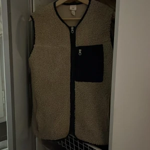 Beige teddyväst  - Säljer en mysig beige fleeceväst från H&M med svarta detaljer. Västen har en dragkedja framtill och bröstficka med dragkedja.