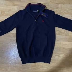Mörkblå tröja från Polo Ralph Lauren - Snygg mörkblå tröja från Polo Ralph Lauren med dragkedja vid halsen och röd logga på bröstet. Tröjan har långa ärmar och ribbade muddar. Perfekt för en stilren look. #g
