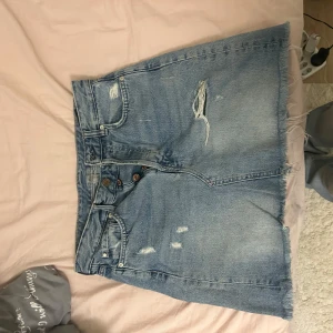 Jeanskjol från Zara - Säljer en cool jeanskjol från Zara i storlek 38. Kjolen har en avslappnad look med slitningar och fransig nederkant. Den är i klassisk blå denim och har knappar framtill. Perfekt för en avslappnad stil!