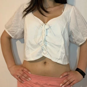 Vit croppad blus med snörning - Säljer en söt vit croppad blus med snörning framtill. Blusen har korta ärmar och en v-ringad design som ger en avslappnad och trendig look. Perfekt för varma dagar!