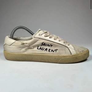 Vita sneakers från Saint Laurent - Snygga vita sneakers från Saint Laurent med handskriven logga på sidan. Skorna har en sliten look med revor och en beige sula. Perfekta för en avslappnad stil. Självklart äkta! Endast dustbag tillkommer!! Kan göra rent skorna innan jag fraktar! 