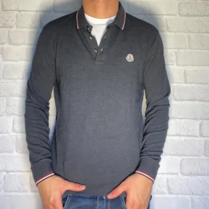 Grå pikétröja från Moncler - Snygg grå pikétröja från Moncler med långa ärmar och klassisk krage. Tröjan har knappar framtill och Monclers logga på bröstet och nfc o scan kod funkar, perfekt för en stilren look.