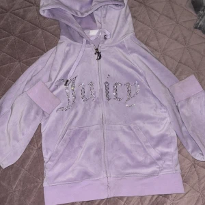 Lila hoodie från Juicy Couture - Säljer en lila hoodie från Juicy Couture med glittrande stenar som bildar texten 'Juicy' på framsidan. Den har dragkedja och huva, samt långa ärmar och fickor framtill. Perfekt för en avslappnad stil med en touch av glamour. Har bara används några få gånger!