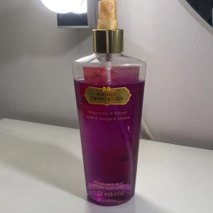 Mango Temptation Victoria secret Fragrance Mist - En fräsch och exotisk doftspray från Victoria's Secret med noter av mango och hibiskus. Flaskan är genomskinlig med en lila vätska och har en gyllene spraypump. Perfekt för att ge en tropisk touch till din dag.