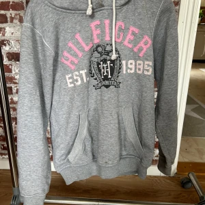 Grå hoodie från Tommy Hilfiger - Säljer en grå hoodie från Tommy Hilfiger med rosa och vita tryckdetaljer. Hoodien har en känguruficka och justerbar dragsko i huvan. Perfekt för en avslappnad stil.(liten i storleken)