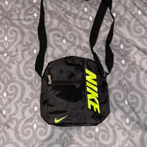 Svart axelväska från Nike - Snygg svart axelväska från Nike med neongul logga. Väskan har en justerbar axelrem och en praktisk dragkedja framtill. Perfekt för att bära med sig det nödvändigaste på språng.