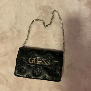 Svart axelväska från Guess - Snygg svart axelväska från Guess med kedjerem och präglad logotyp. Kan ha den på olika sätt , knappt andvänd 