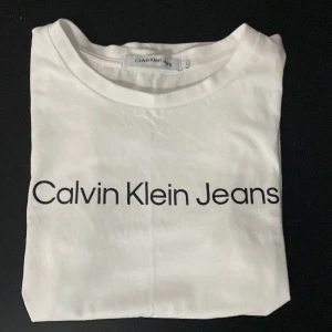 Vit t-shirt från Calvin Klein Jeans - Säljer en stilren vit t-shirt från Calvin Klein Jeans med svart logotyptryck på framsidan. Perfekt för en casual look. T-shirten är tillverkad i mjukt bomullsmaterial och har en klassisk passform. Använt 1 gång.
