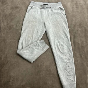 Grå mjukisbyxor från Tommy Hilfiger ( Äkta ) Köpt från Johnells - Snygga grå mjukisbyxor från Tommy Hilfiger med snörning i midjan och ribbade muddar vid bensluten. Byxorna har en diskret logga längs benet och en bakficka. Perfekta för en avslappnad stil.