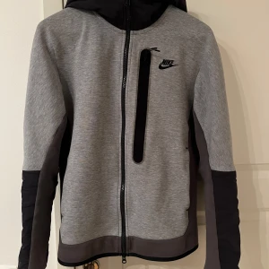 Grå och svart jacka från Nike - Snygg grå och svart jacka från Nike med dragkedja och huva. Perfekt för kyligare dagar med sin stilrena design och praktiska fickor. Jackan har långa ärmar och en normal passform.