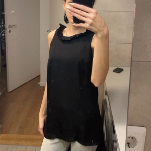 Svart ärmlös topp med volangdetalj - Snygg svart ärmlös topp med volangdetalj vid halsringningen. Perfekt för en stilren look. Och kanske en utekväll. (Lite genomskinlig och tunn så man får ha nått under)