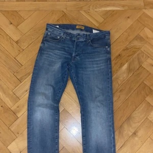 Blå jeans från Jack & Jones - Snygga blå jeans från Jack & Jones med klassisk femficksdesign och en bekväm passform. De har en dragkedja och knapp framtill samt en läderpatch med logotyp på baksidan. Perfekta för en avslappnad stil.