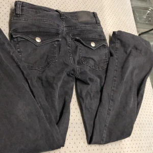 Grå jeansbyxor med fickdetaljer - Snygga grå jeansbyxor med två bakfickor som har knappdetaljer. Perfekta för en casual look med en t-shirt eller en snygg blus. Byxorna har en klassisk design med en modern touch.