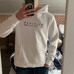 Tommy hilfiger hoodie vit  - Säljer en stilren hilfiger hoodie med texten ”Hilfiger New York” på bröstet. Tröjan har en klassisk design med känguruficka. Knappt använd, skick 10/10.