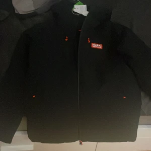 Svart skidjacka från Huski Wear - Säljer en helt ny svart jacka från Huski Wear då jag fick den av min farbrors fru som jobbar där men jag har redan en skidjacka med dragkedja och quiltad insida. Jackan har en stilren design med röda detaljer och är perfekt för kyligare väder. Den är tillverkad av återvunna material vilket gör den till ett miljövänligt val.