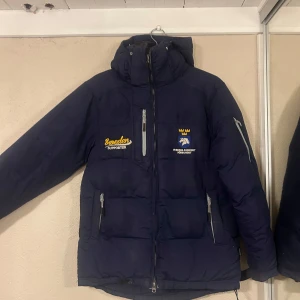 Blå dunjacka med broderad logga - Säljer en blå dunjacka med broderad logga på bröstet. Jackan har dragkedja och flera praktiska fickor. Perfekt för kalla dagar med sin varma och skyddande design. Funkar parfekt till stallet på kalla kvällar och på vinter. Stolek XS men jag har M och den funkar super bra (den kommer nytvättad 