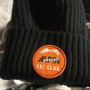 Svart ribbad mössa från TrendEagle - Snygg svart ribbad mössa från alpin garaget med ett broderat märke i orange och vitt framtill. Perfekt för kyliga dagar och en stilren look. Den passar dock bättre på personer med lite större huvud.