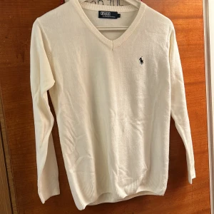 Vit tröja från Ralph Lauren - Säljer en stilren vit tröja från Ralph Lauren med v-ringning och det klassiska logotypbroderiet på bröstet. Tröjan har långa ärmar och ribbade kanter för en bekväm passform. Perfekt för en elegant och avslappnad look.