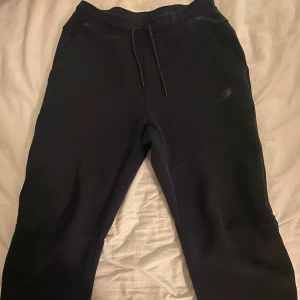 Svarta mjukisbyxor från Nike - Säljer ett par svarta mjukisbyxor från Nike med snörning i midjan och dragkedja på fickorna. Perfekta för en avslappnad stil eller träning. Byxorna har en diskret logga på benet.