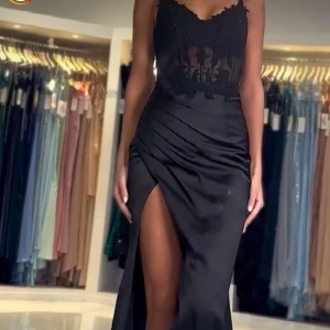 Svart långklänning med spetsdetaljer, bal klänning - Elegant svart långklänning med spetsdetaljer och transparent överdel. Klänningen har en djup slits och smala axelband, vilket ger en sofistikerad look. Perfekt för en glamorös bal.