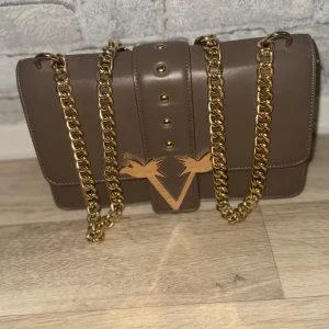 Brun axelväska med guldkedja - Snygg brun axelväska med guldkedja och dekorativa nitar. Väskan har ett elegant märke framtill och flera fack inuti för praktisk förvaring. Perfekt för att ge din outfit en lyxig touch.