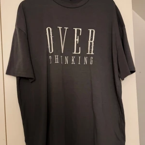 Svart t-shirt med text från Shein - Säljer en svart t-shirt från Shein med trycket 'OVERTHINKING' i vitt på framsidan. T-shirten har en normal passform och korta ärmar, perfekt för en avslappnad stil.