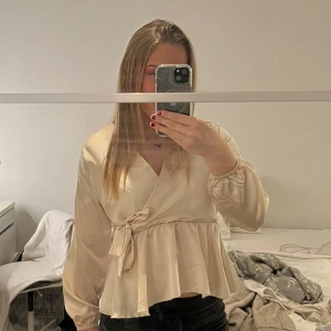 Beige omlottblus med knytband - Säljer en elegant beige omlottblus med knytband i midjan. Blusen har långa ärmar och en volangdetalj nedtill som ger en feminin touch. Perfekt för en stilren look.