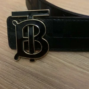 Svart och beige bälte med rutigt mönster - Snyggt burberry bälte med ett stort, stilrent spänne i svart och guld. Ena sidan är svart med diskret rutmönster och den andra är beige med klassiskt rutigt mönster. Perfekt för att ge din outfit en extra touch.