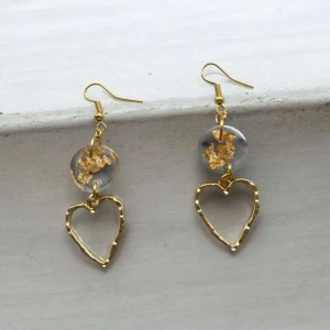 Guldiga hjärtformade örhängen - Eleganta örhängen med hjärtformad design i guld. De har en klar pärla med guldflingor ovanför hjärtat, vilket ger en unik och stilren look. Perfekta för att lägga till en touch av glamour till din outfit.