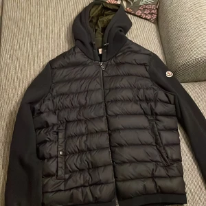 Svart/ mörkblå cardigan från Moncler - Säljer en snygg svart pufferjacka från Moncler med huva och dragkedja. Jackan har en quiltad design på framsidan och ett Moncler-märke på ärmen. Perfekt för sommar kvällar, höst och vår dagar med sin stilrena look och bekväma passform. Inga disfunktioner. Pris kan förhandlas, om du har någon fråga är de bara och kontakta mig. Kan gå med på byten