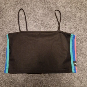 Svart tubtopp från Minga London - Säljer en cool svart tubtopp från Minga London med färgglada ränder i blått, rosa, rött och gult på sidorna. Toppen har smala axelband och en dragkedja bak. Perfekt för en trendig look!