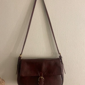 Vinröd axelväska i skinn - Snygg vinröd axelväska i skinn med justerbar axelrem och ett elegant spänne framtill. Väskan har en praktisk innerficka med dragkedja för extra säkerhet. Perfekt för att bära dina dagliga nödvändigheter med stil.